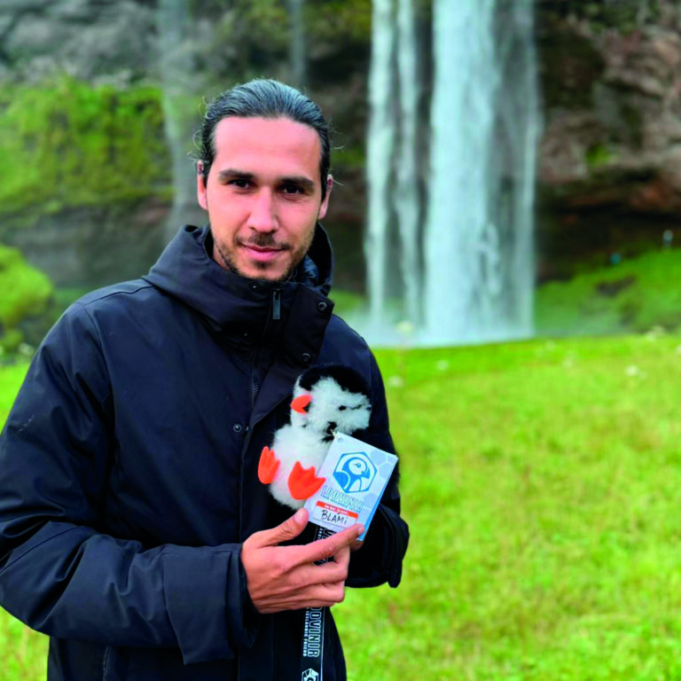 Frank holding his adopted LOÐVINIR puffin plush named Lundi in front of Skógafoss waterfall in Iceland
